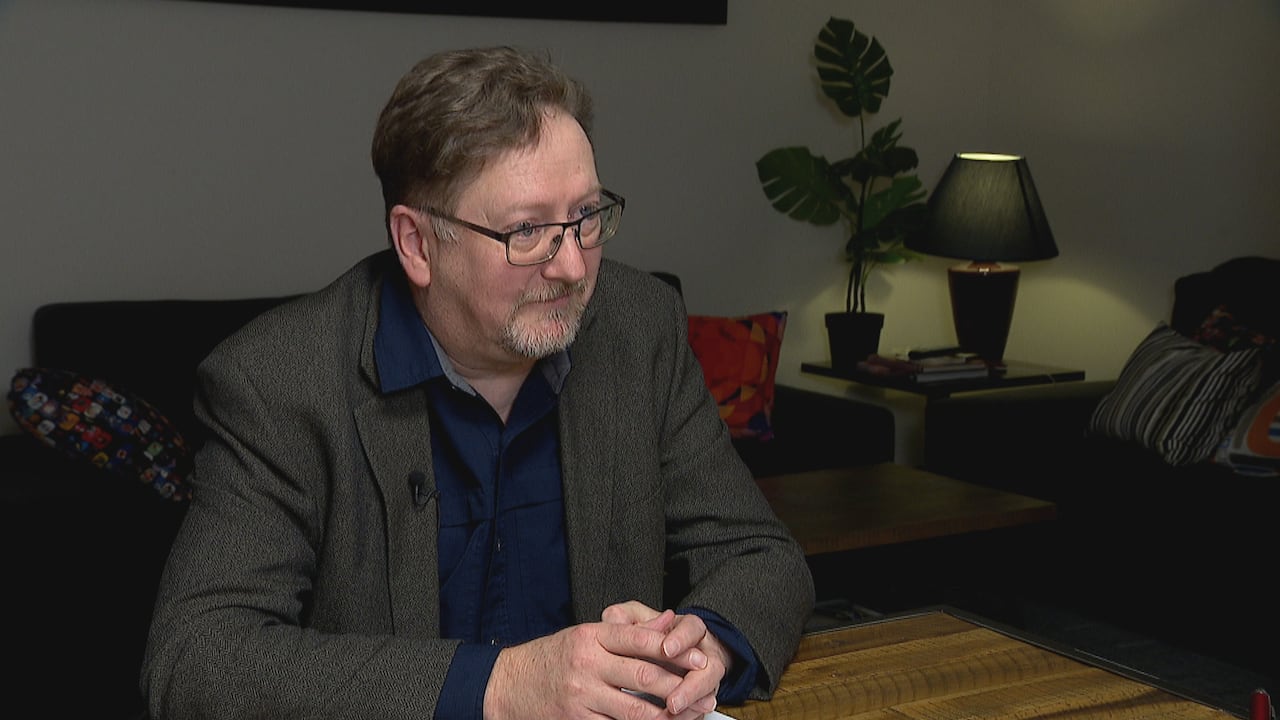 A man wearing a grey jacket and dark blue shirt sits at a table with his hands folded.