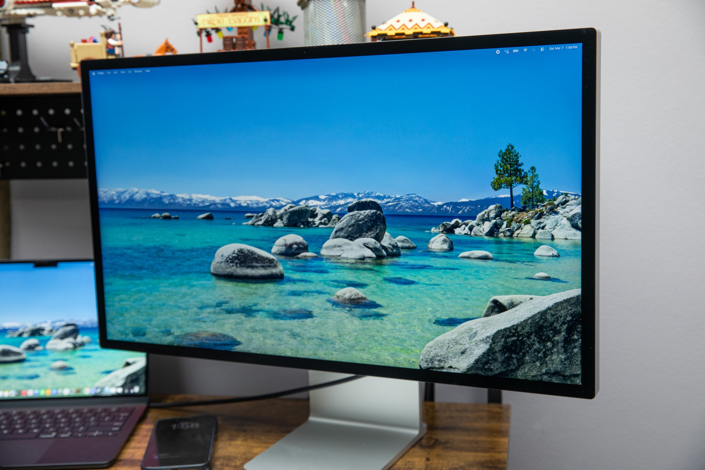 The Apple Studio Display XDR displaying a water landscape on a wooden desk next to a Macbook.