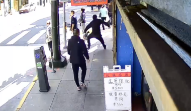 A woman in a burgundy blazer, white top, and black pants stands confidently outside a modern building with large windows reflecting trees.