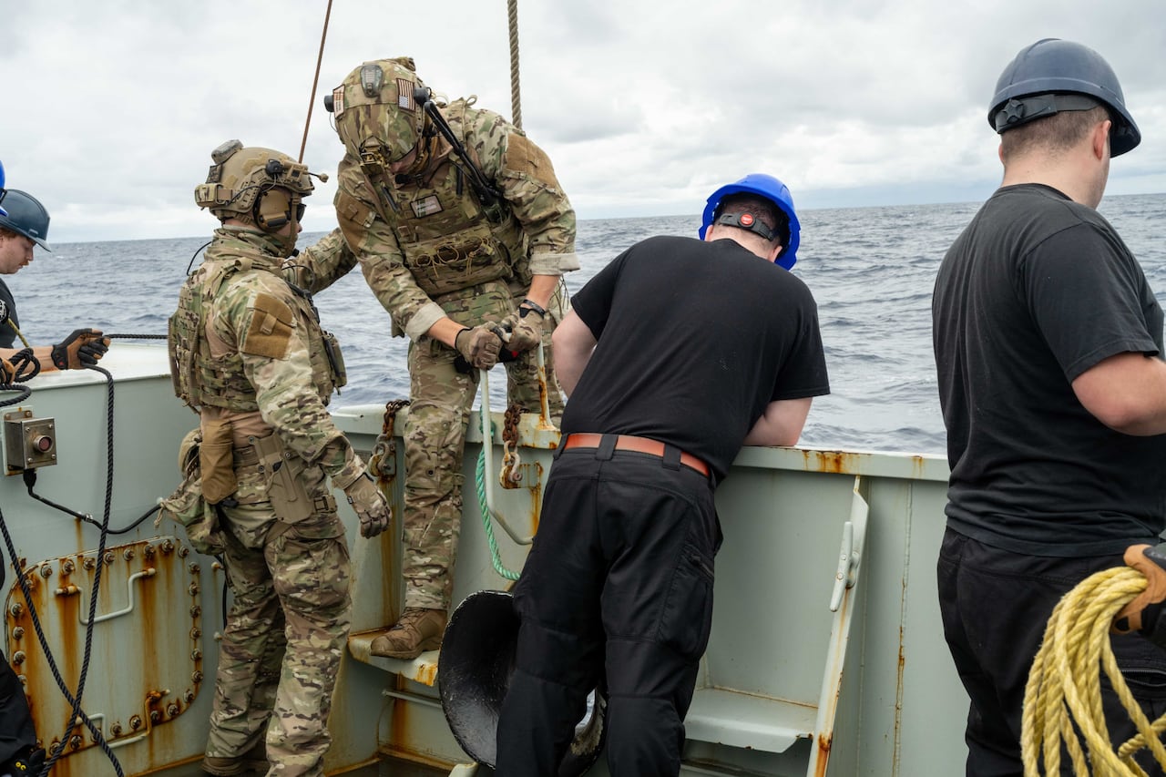 Canadian Navy sailors help a USCG officer in camp to climb over the gunwhale of their ship as he attaches a safety line to himself.