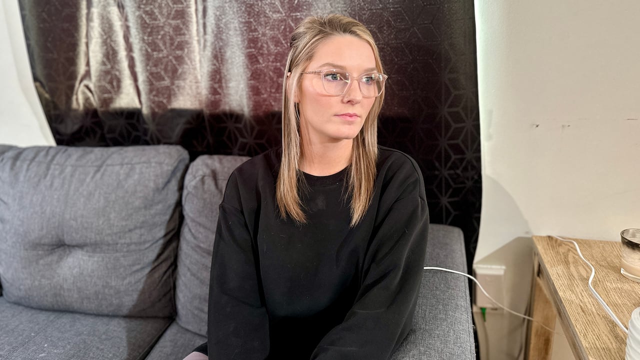 A woman wwearing glasses with short blonde hair sits on a sofa and stares off to the side.