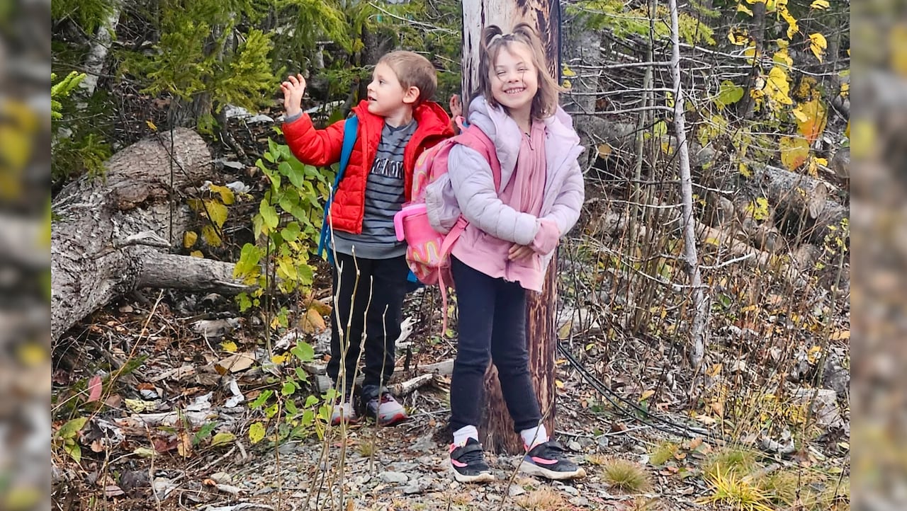 Two children, a boy and a girl, standing in front of woods with backpacks on.