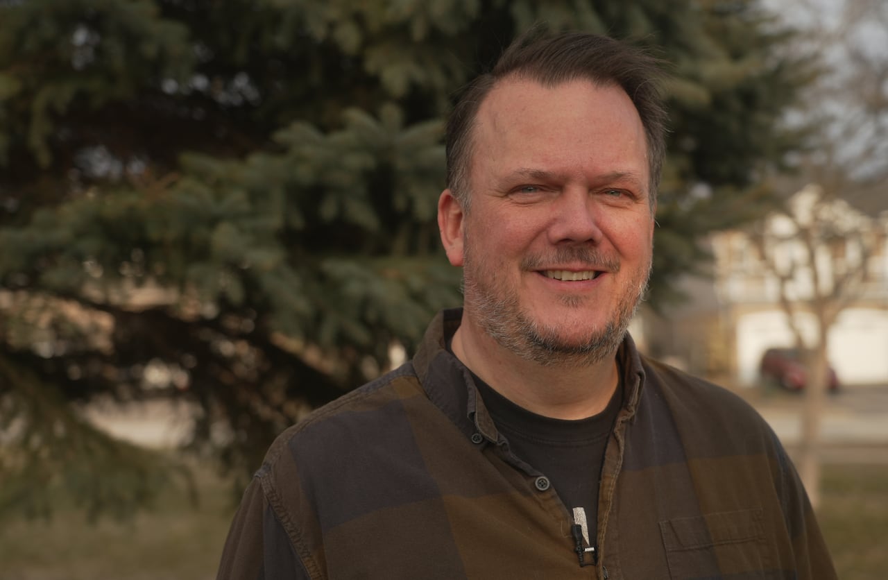 A man with a gray beard smiles while standing in front of an evergreen tree.