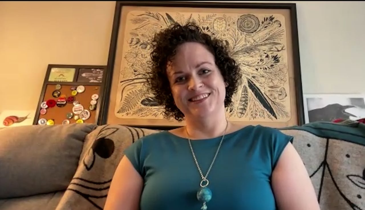 A woman with short, curly dark hair wearing a blue short-sleeve top and necklace. 