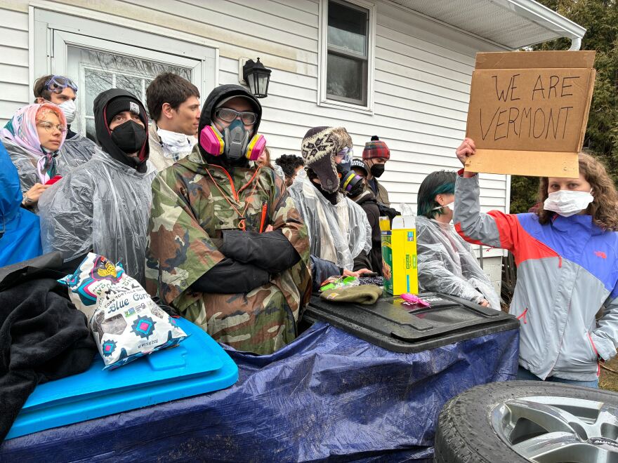People bundled in jackets and wearing medical masks and respirators stand in front of a building. One holds a sign that reads "we are Vermont"