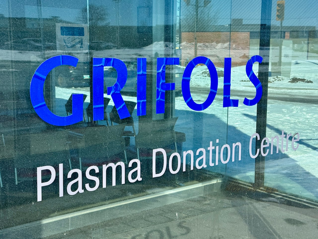 A sign saying Grifols appears on the glass exterior wall of a building with snow seen on the ground near the building.