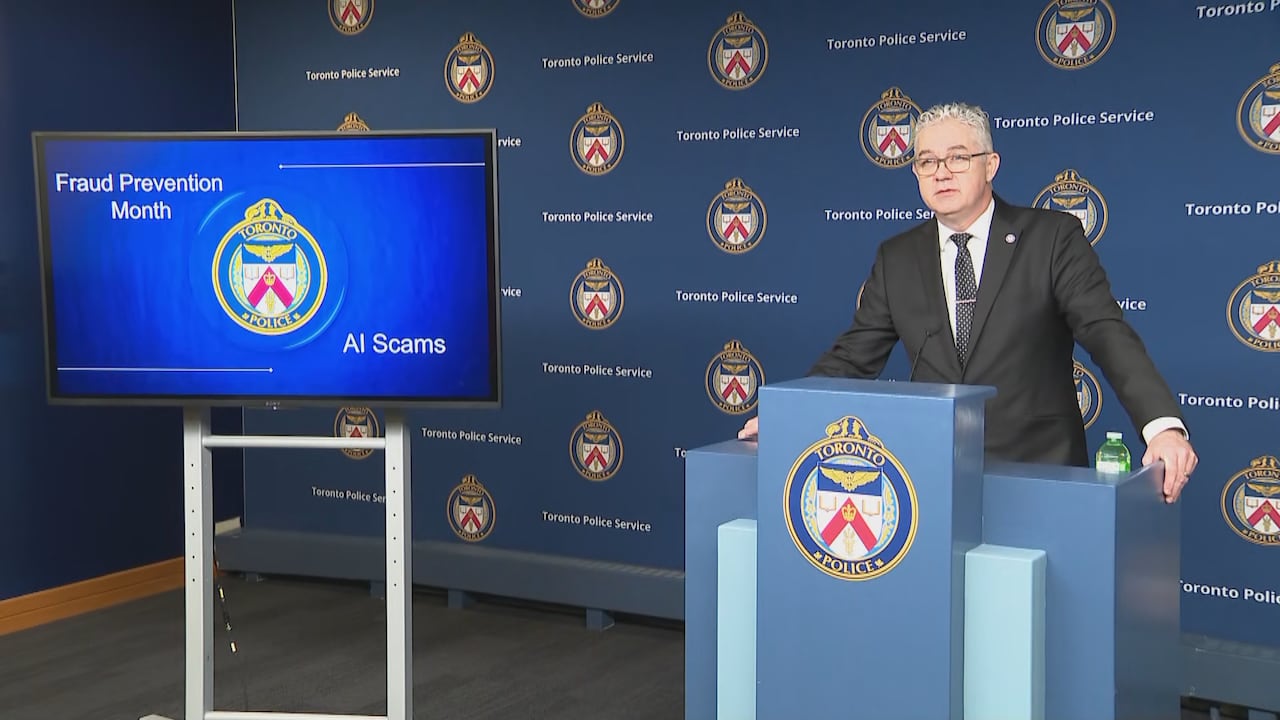 Toronto Police Det. David Coffey stands behind a podium in front of a blue wall TPS background, next to a TV screen that reads fraud prevention month.