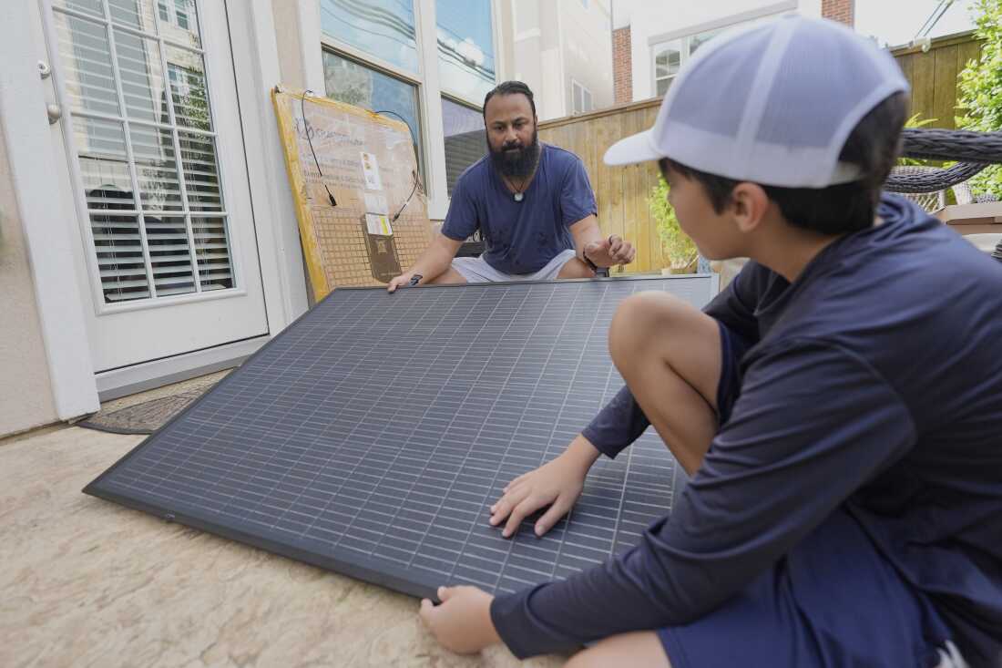 Squatting near the ground outdoors, Bhavin Misra and his son, Rumi, hold opposite sides of a black, square solar panel with a grid pattern on it while assembling a plug-in solar kit at their home in Houston.