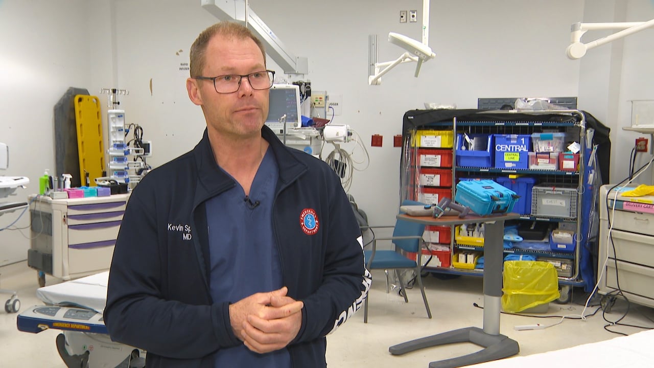 Dr. Spencer stands in a room in the Dartmouth General Hospital with a bed and medical supplies behind him. 