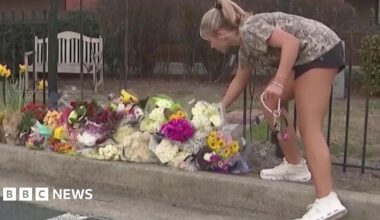 A student in shorts and a T-shirt places flowers on a pile of bouquets at a sidewalk memorial