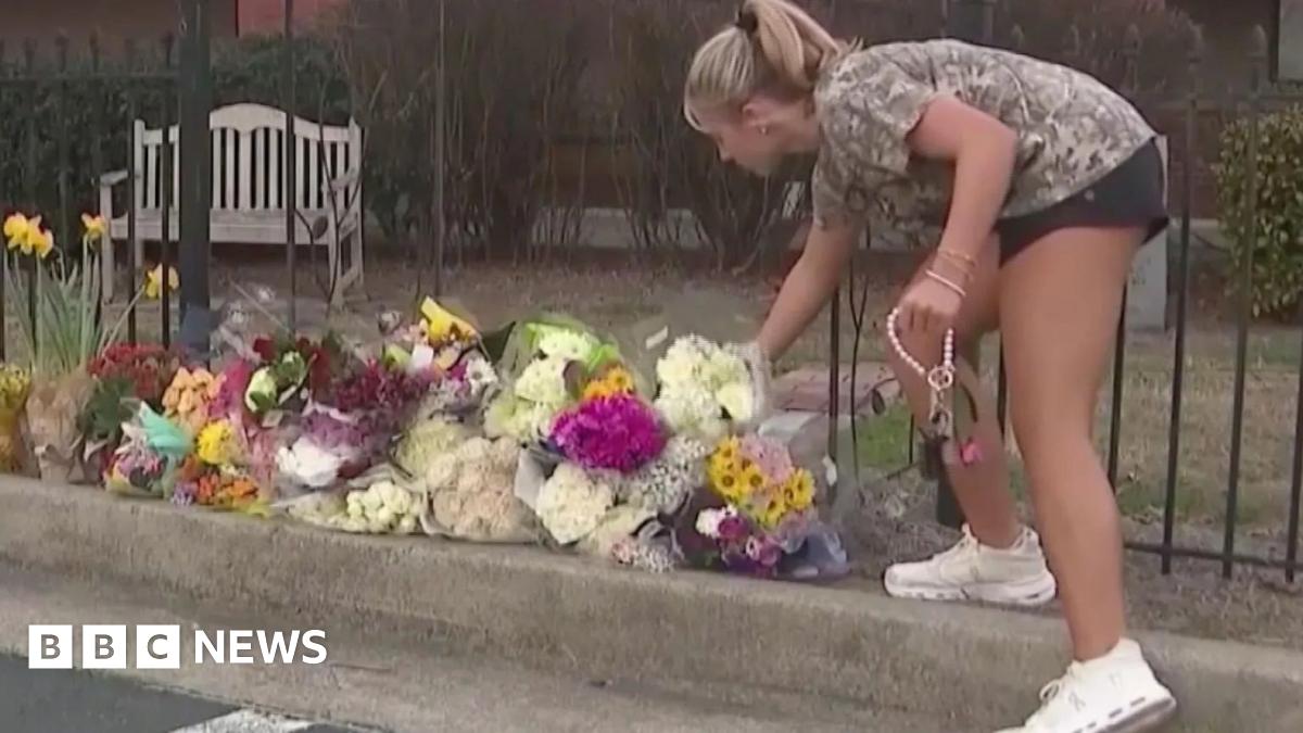 A student in shorts and a T-shirt places flowers on a pile of bouquets at a sidewalk memorial