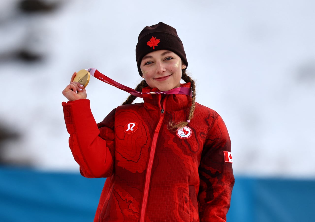 A female Paralympic skier representing Canada smiles while holding up a gold medal that hangs around her neck.