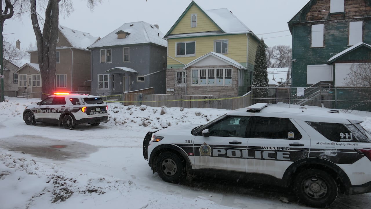 Police cruisers, one with its lights on, are parked on a snowy street. A house in the background has yellow tape around the front.