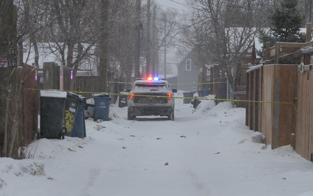 A police cruiser is parked in a snowy back alley with yellow caution tape blocking the lane.