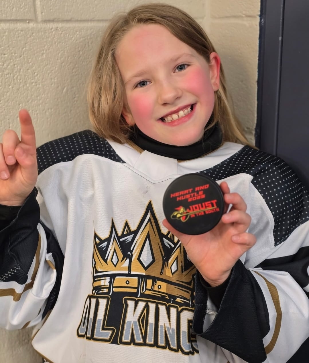 A young blonde girl looks at the camera holding a puck and number one hand sign.