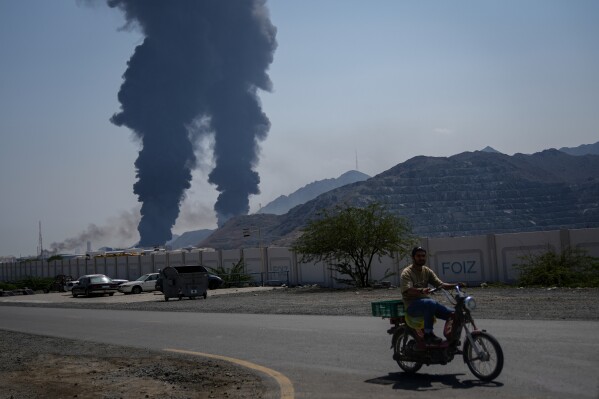 A man rides a bike as fires and plumes of smoke rise after debris from an intercepted Iranian drone struck an oil facility, according to authorities, in Fujairah, United Arab Emirates, Saturday, March 14, 2026. (AP Photo/Altaf Qadri)