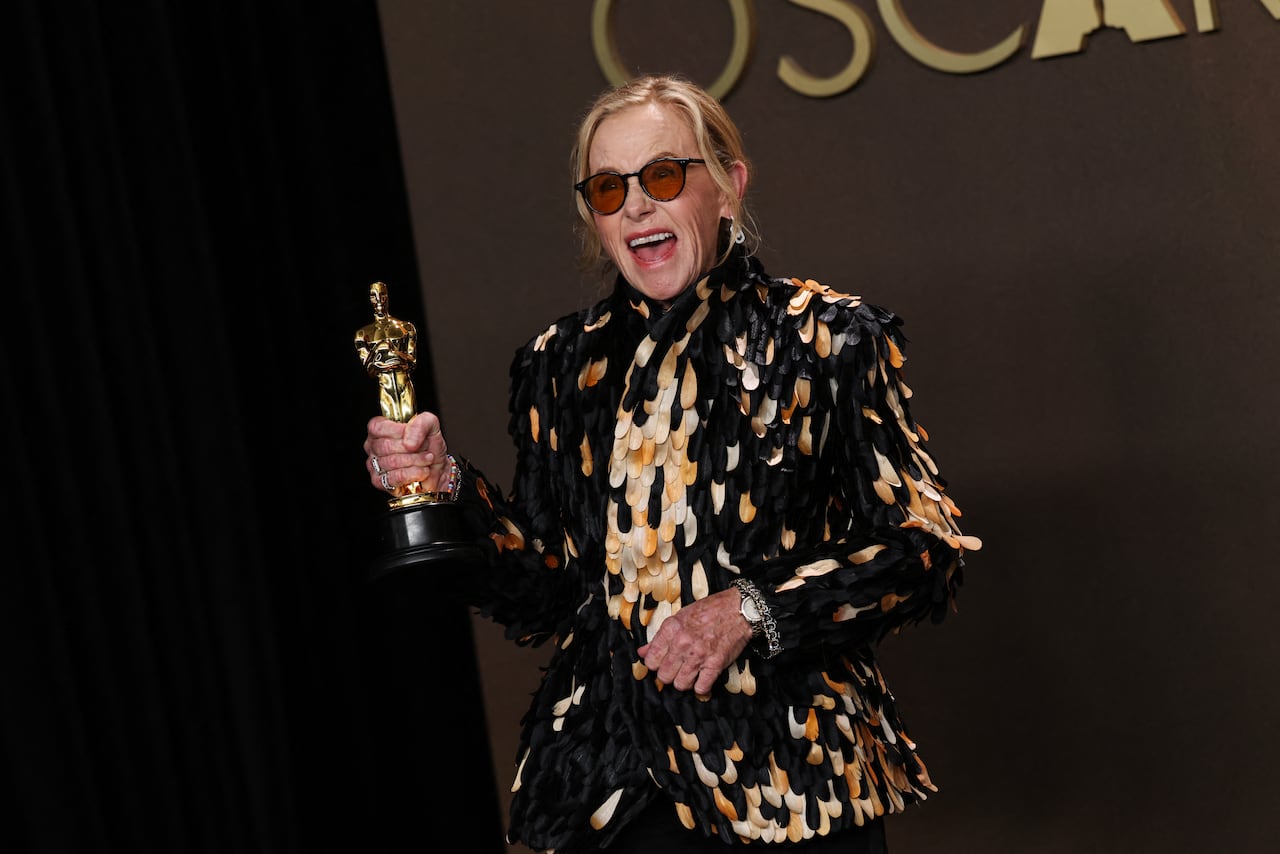A woman wearing sunglasses a jacket covered in black, white and brown feather paillettes holds up a gold Oscar statuette as she laughs while posing for a photo.