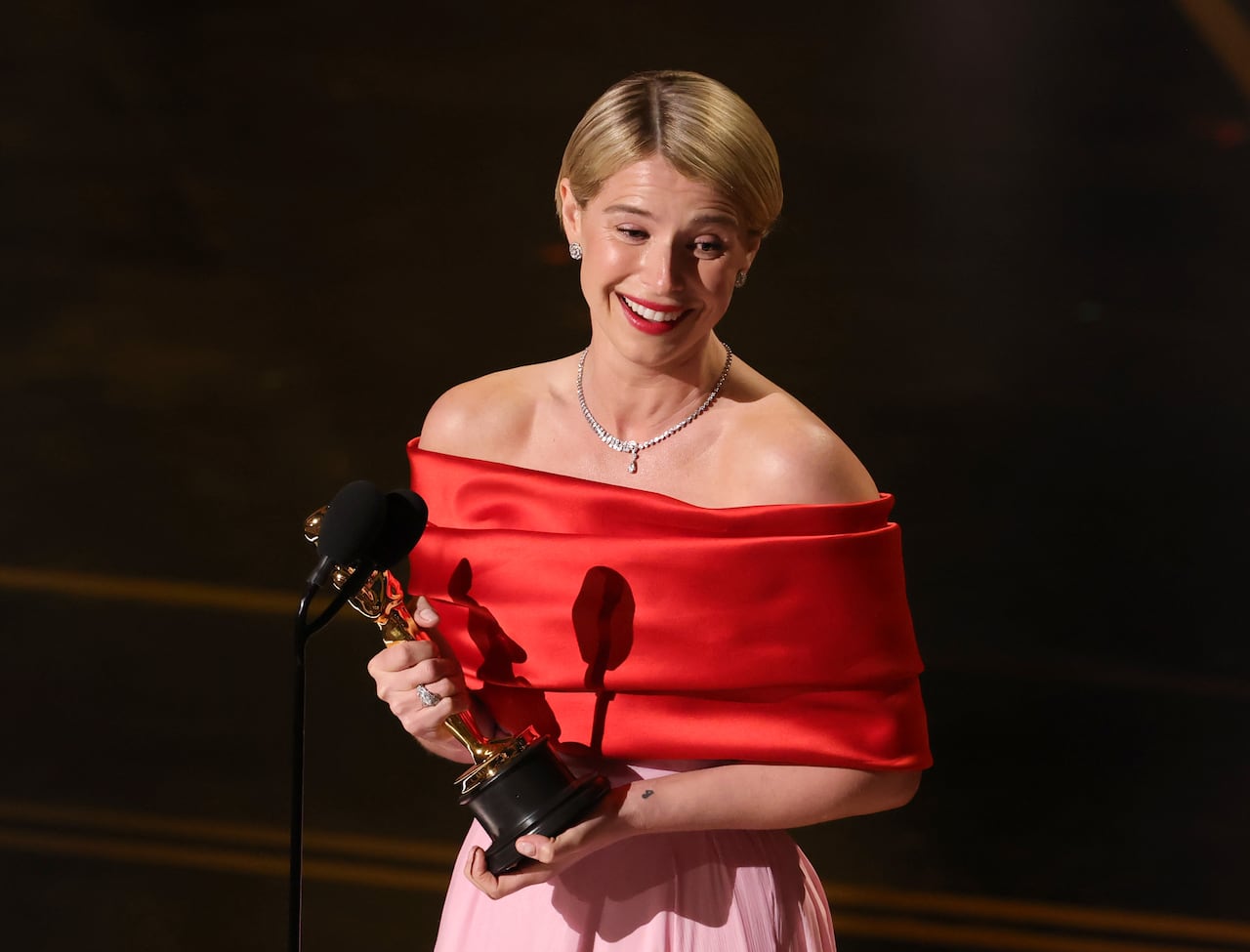 A woman, with short blond hair and wearing a red and pink off-the-shoulder gown, holds a gold Oscar statuette in her hands as she smiles on stage.