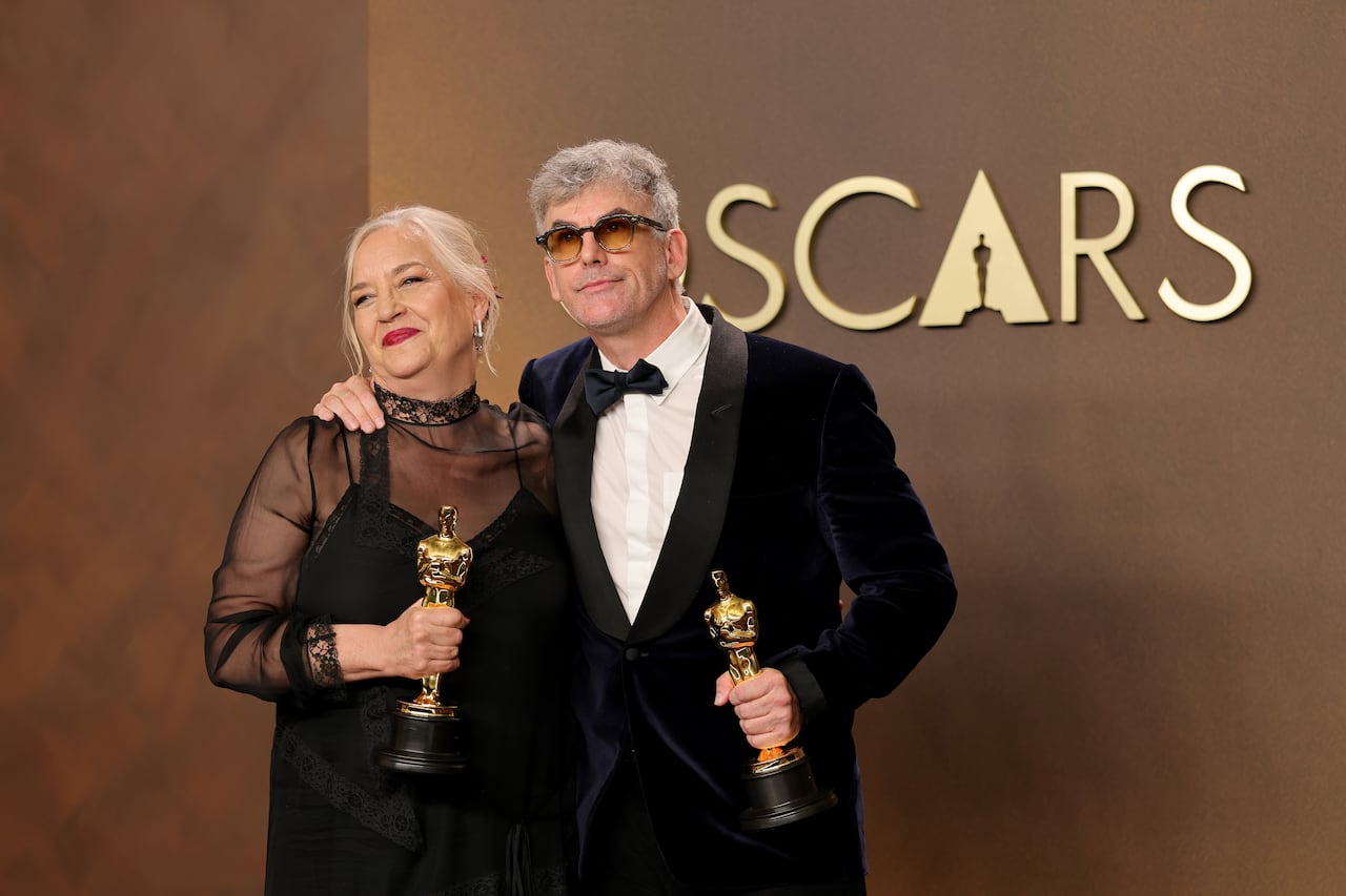 A woman and a man, both wearing black formal outfits, stand next to each other while smiling and holding gold Oscar statuettes in their hands.