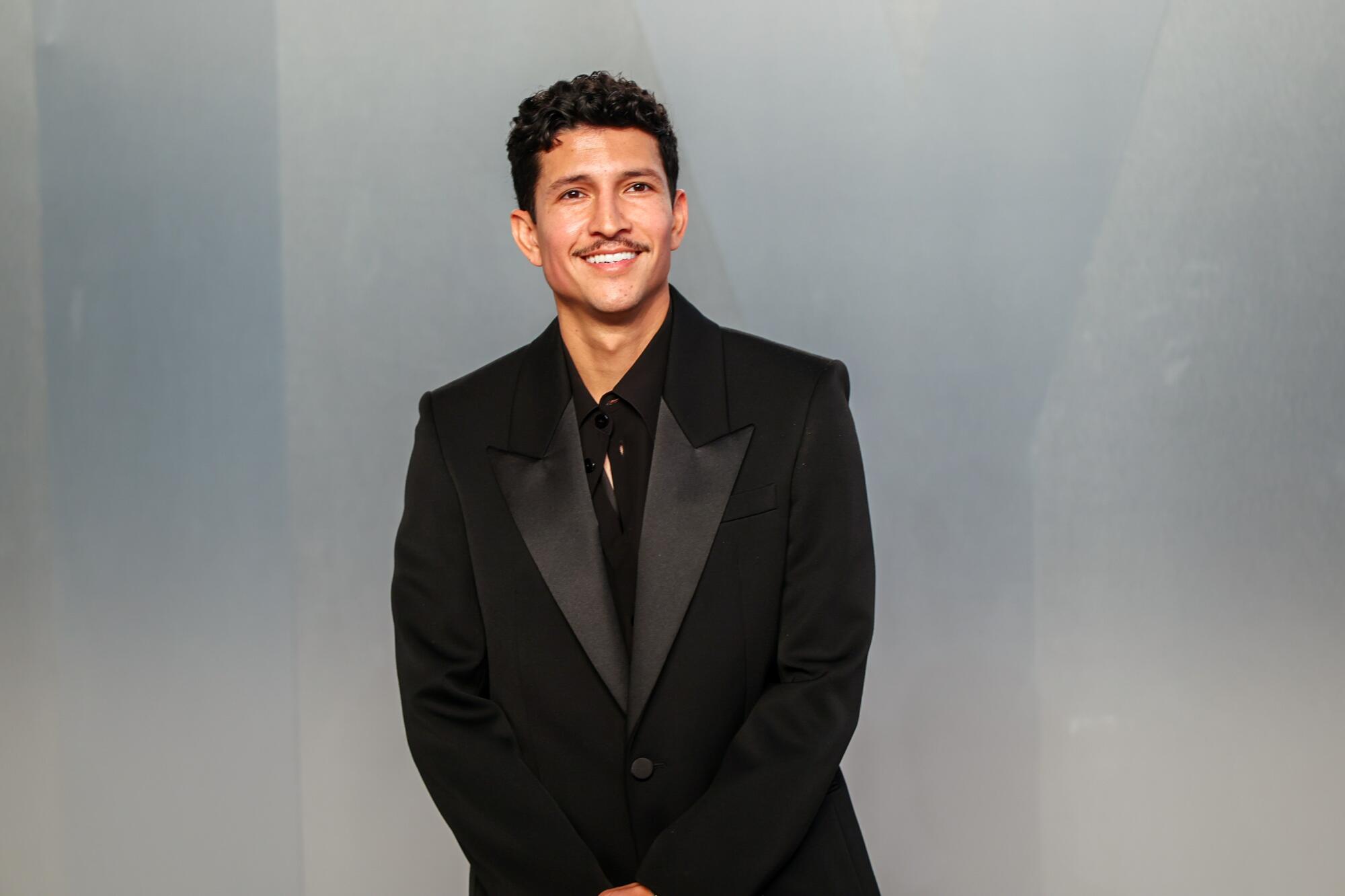 Danny Ramirez, in a black suit, smiles on the red carpet. 