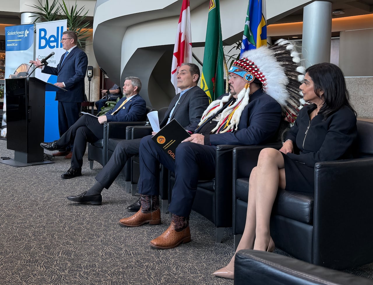 A man in a suit speaks at a lectern next to four people sitting in chairs, including one wearing a First Nation's headdress.