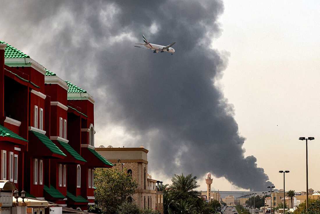 An Emirates aircraft prepares for landing as a smoke plume rises from an ongoing fire near Dubai International Airport in Dubai on March 16, 2026.
