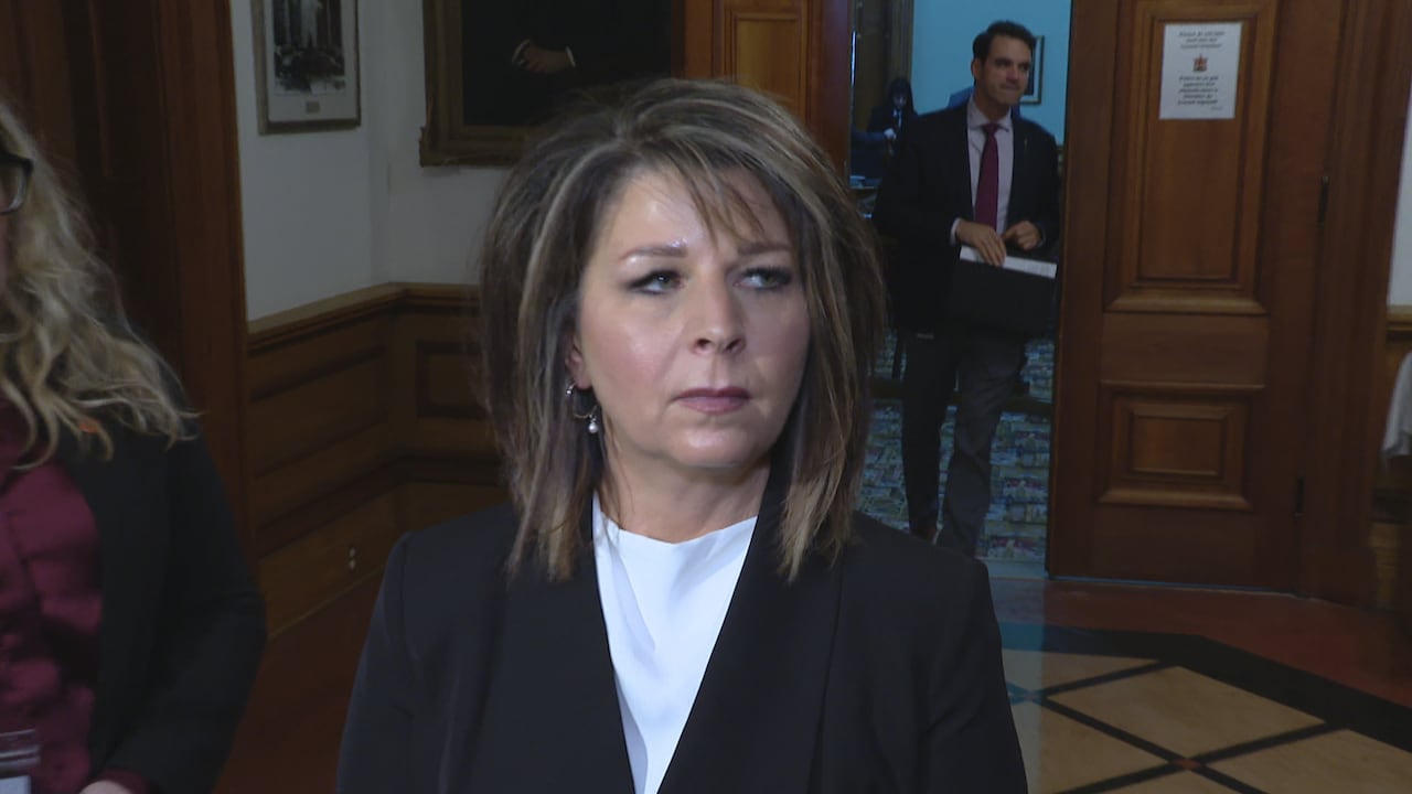 A woman with dark hair and highlights standing outside of a committee room looks to the right.