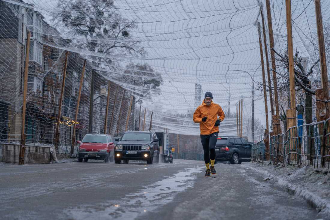A man jogs in the street beneath nylon netting strung over the road. 