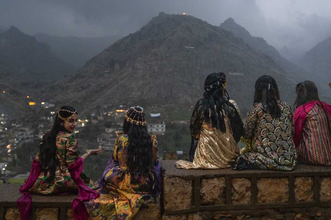 Five teen girls sit on a low wall to get a good vantage point of people walking up the mountains with flaming torches and fireworks. They are wearing colorful gowns and some have gold headpieces over their long hair.