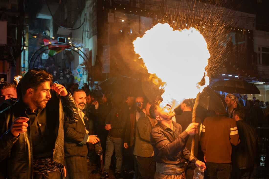 A man spits fire on a torch as other people walk close by.