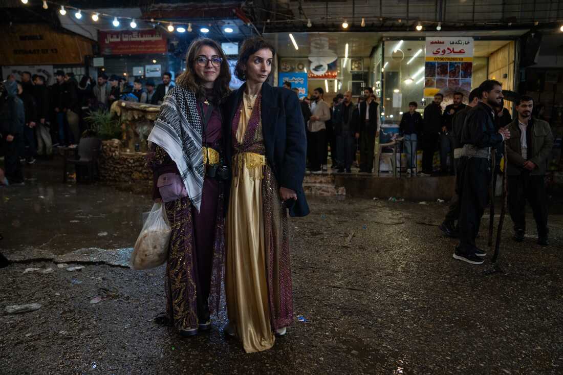 Avjin Yazgan, 21, stands with her mother Pelda Yazgan, 43, are visit Akre to celebrate the holiday. Avjin is wearing a maroon dress and a keffiyeh. She is holding a bag of flatbread. Pelda is wearing a gold and maroon dress with a suit jacket over top. 