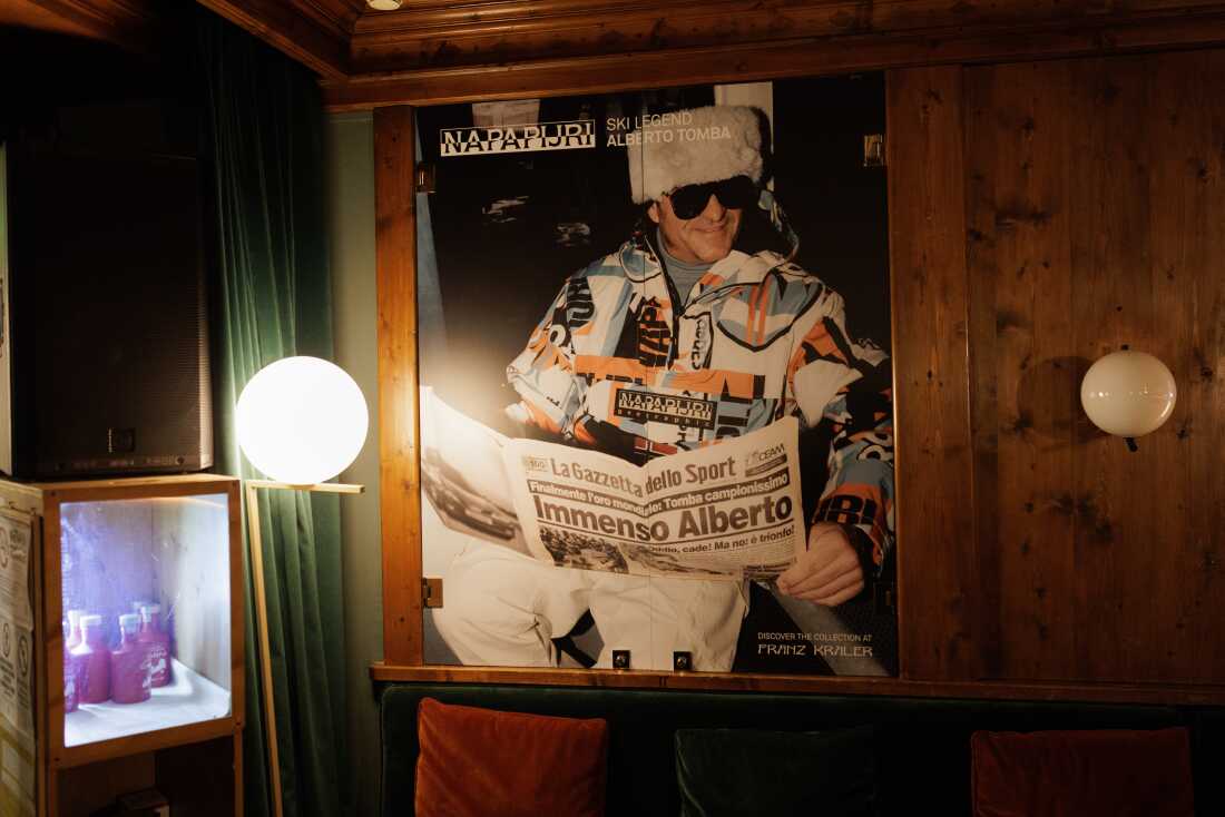 A large-scale photograph of Italian skiing legend Alberto Tomba, wearing a traditional fur hat and reading a sports newspaper with the headline "Immenso Alberto," is displayed in a wood-paneled interior. 