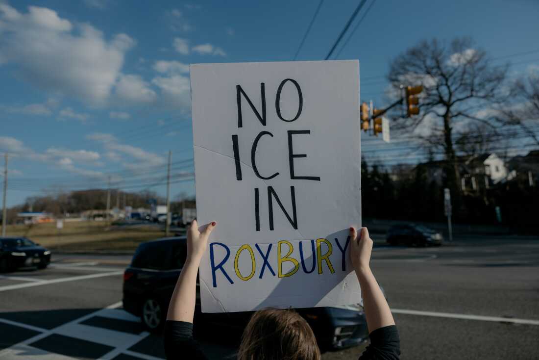 A protester standing along a street holds up a sign that says in all capital letters: "NO ICE IN ROXBURY."