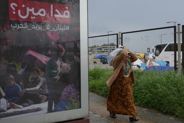 A displaced woman who fled Israeli airstrikes in southern Lebanon, carries her belonging as she moves to a better spot to shelter from the rain, past an Arabic anti-war poster that reads, "Sacrificing for whom? Lebanon does not need war," in Beirut, Saturday, March 21, 2026.(AP Photo/Hussein Malla)
