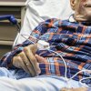 An older man lies in a hospital bed with guardrails. He is wearing a blue and red plaid shirt and having his pulse oxygen level measured with a device on his finger. His face is only partially visible.