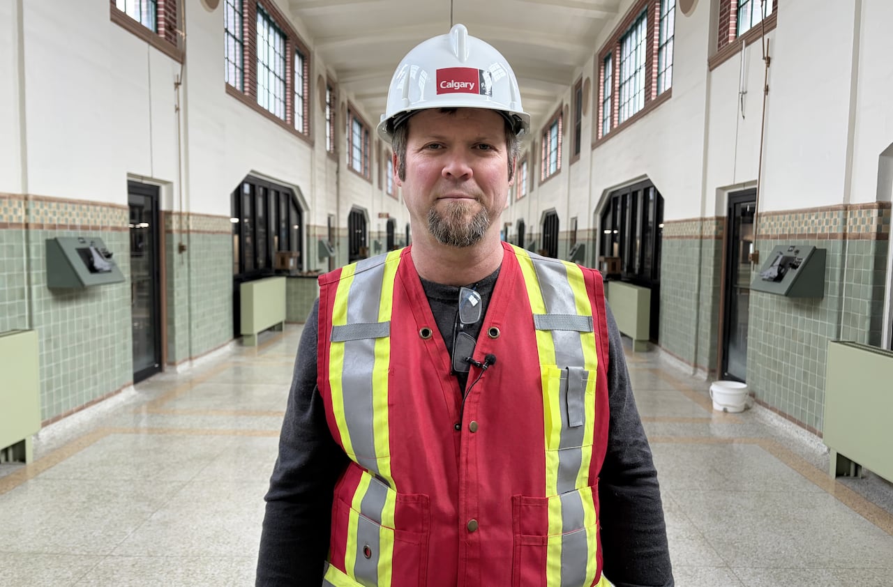 A man wearing a safety vest and hard hat with the City of Calgary logo stands in the middle of a long hallway.