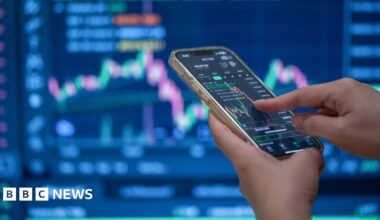 A trader's hands using their phone to make a trade in front of a trading screen in soft focus