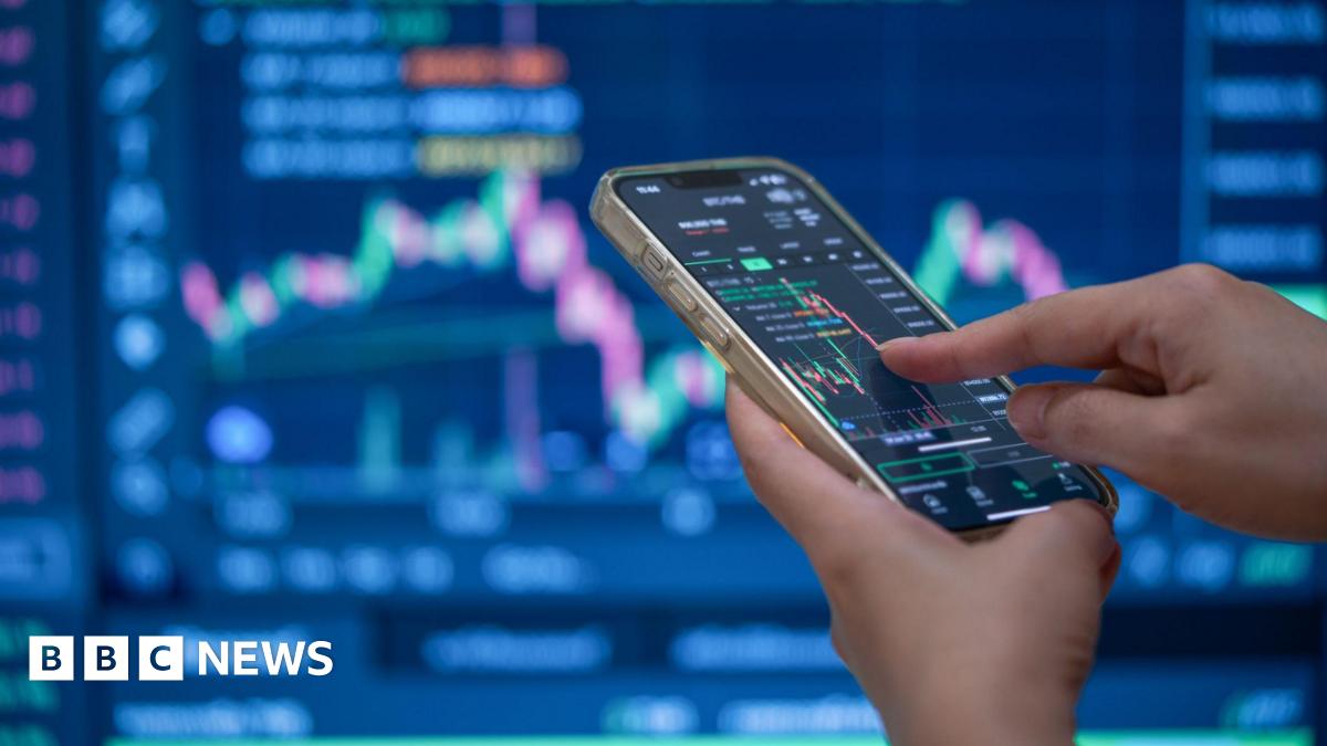 A trader's hands using their phone to make a trade in front of a trading screen in soft focus