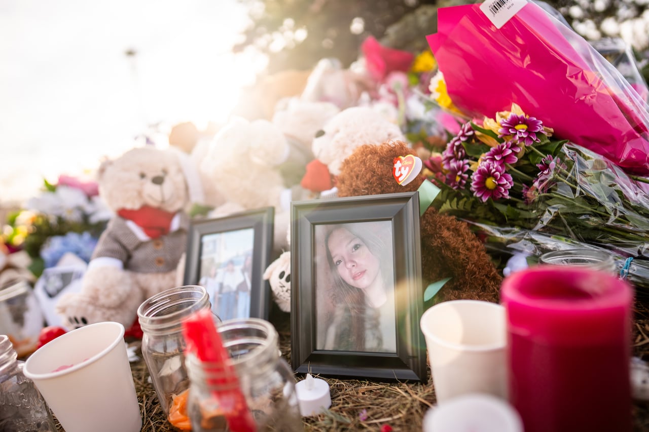 A closeup of a makeshift memorial with pictures of children, teddy bears, vigil candles and flowers on the ground, with sun rays up above.