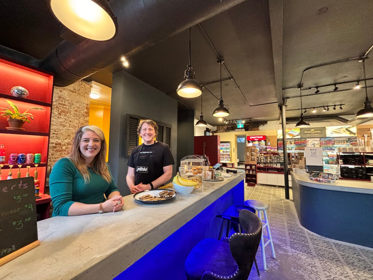 A white woman with blonde and brown hair and a white man with brown hair stand behind a counter that has a plate of cookies on it in a store.