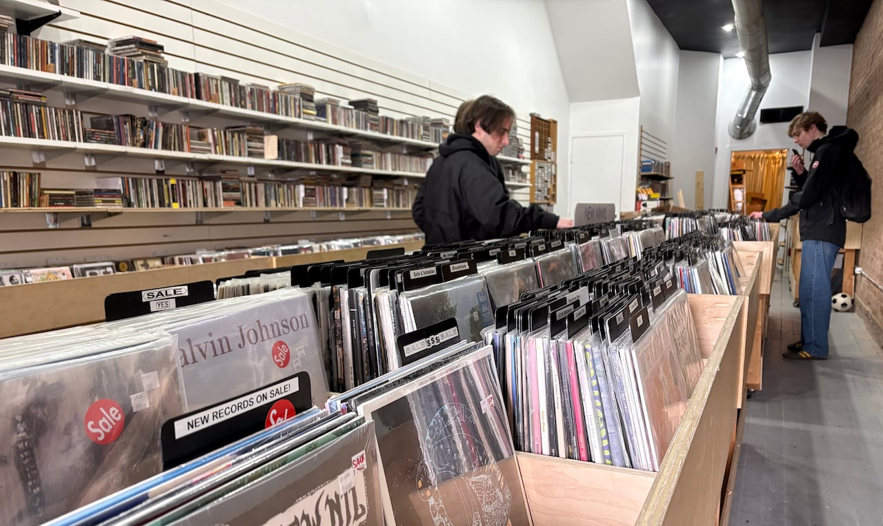 Two men leaf through records in a record store, which has wooden racks full of vinyls. 