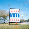 A "VOTE" sign stands near a polling center in Austin, Texas. The sign says "VOTE" in blue capital letters, and rows of five red stars are above and below the word.