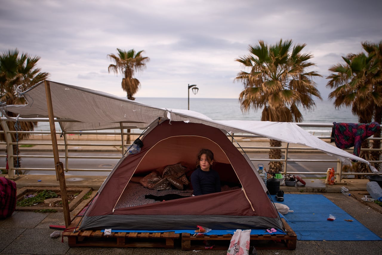 A small girl sits inside a tent that has been erected in a public place, near a shoreline.