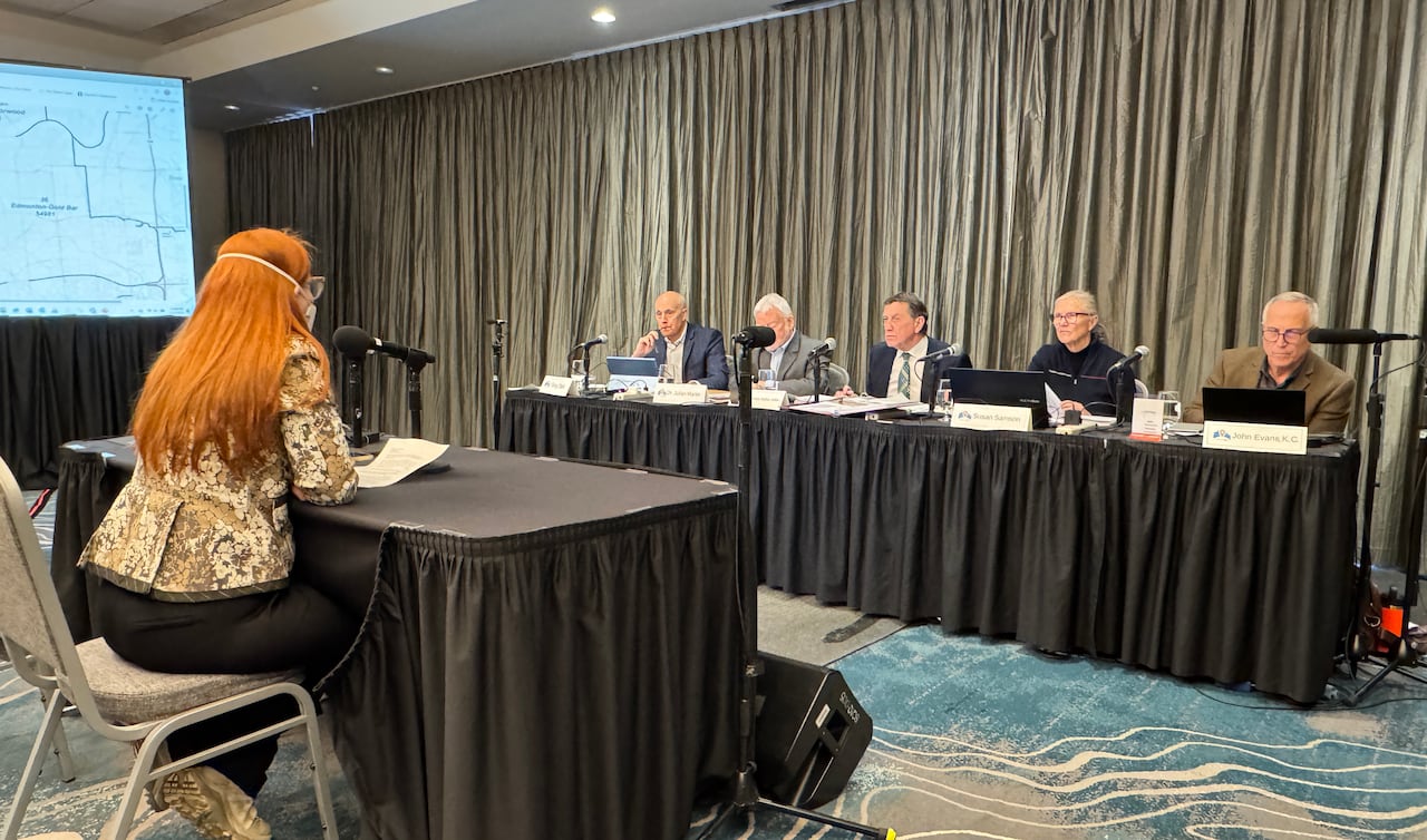 Five people sit behind a long table with mics in front of their faces. A woman sits at a table across from them.
