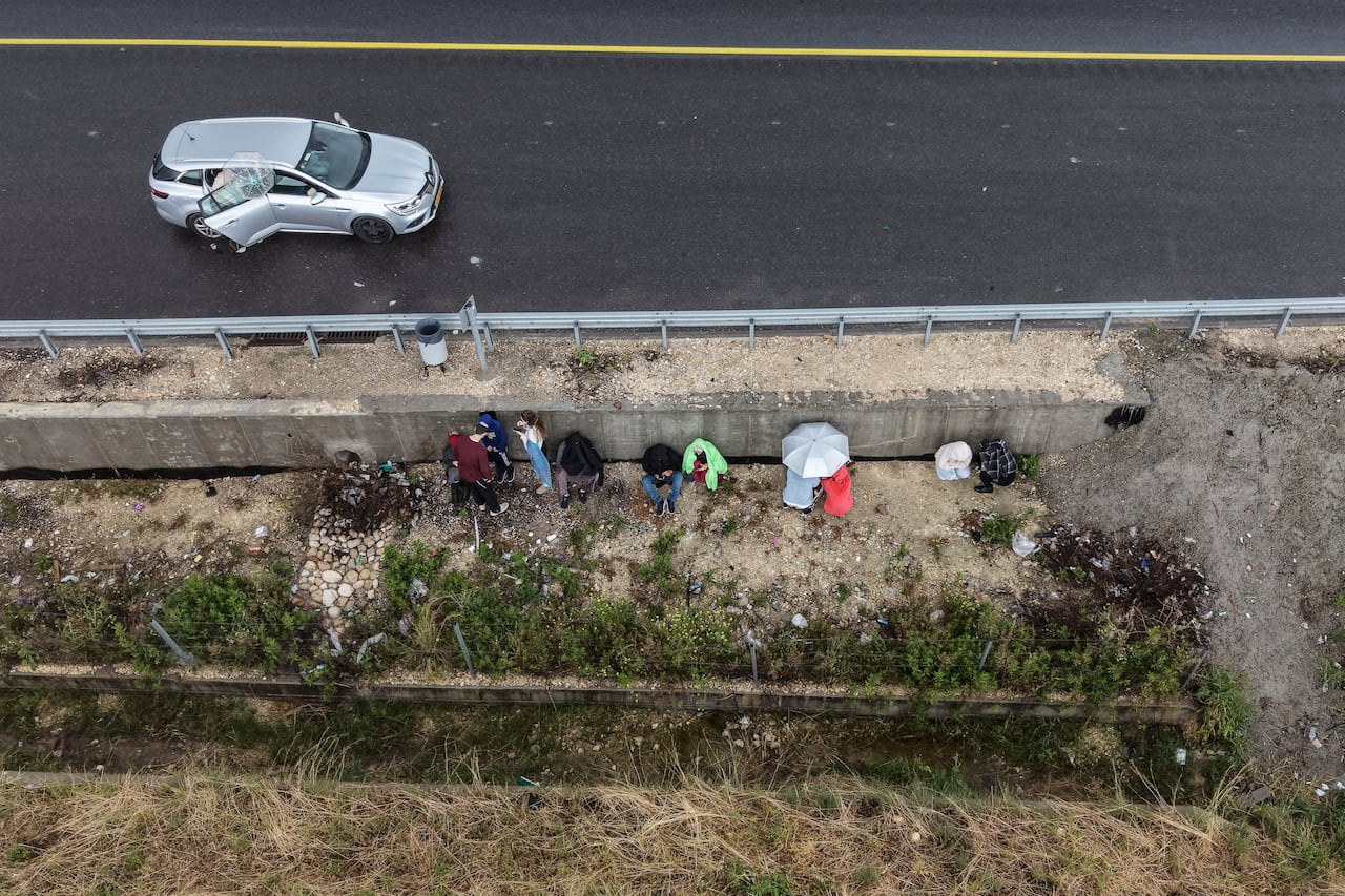 An overhead drone photo show people taking shelter at the side of a road. An abandoned car sits by the side of a highway.