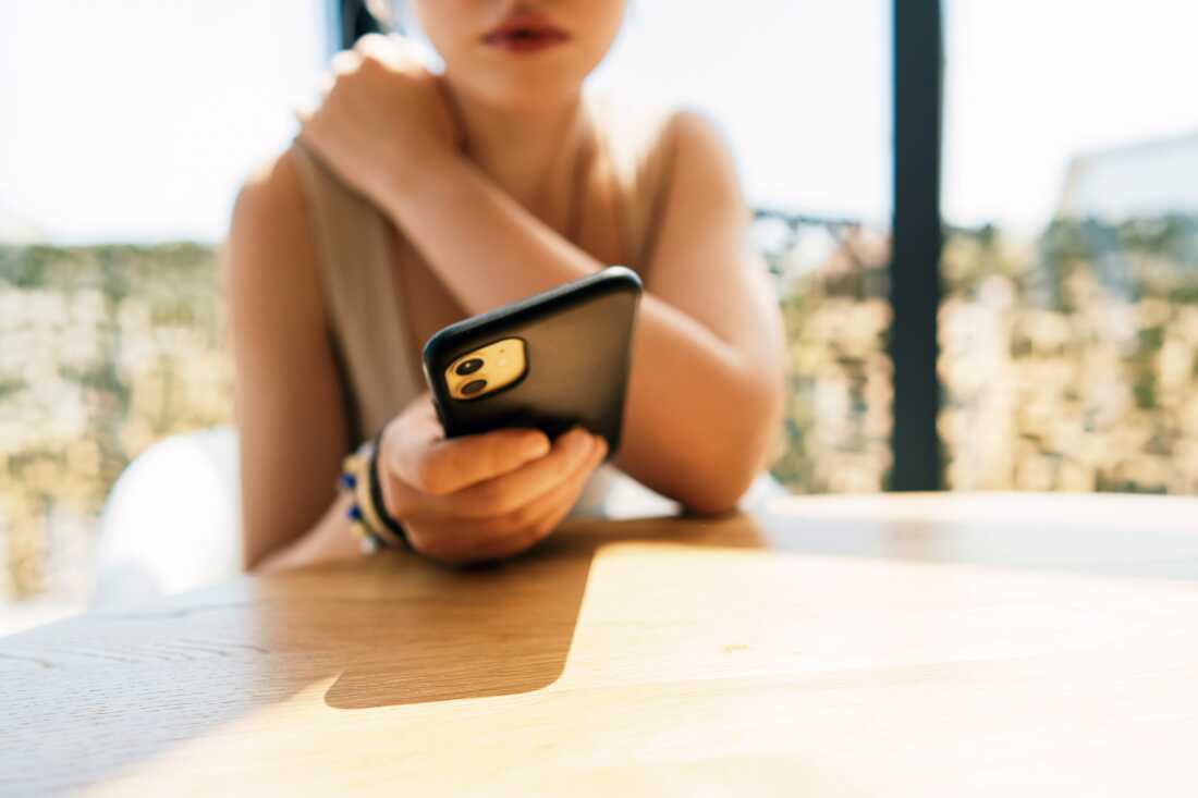 A young woman sits at a sunlit table, holding a smartphone with a soft focus background. The scene conveys casual connectivity, focus on the device, and a relaxed moment of everyday life.
