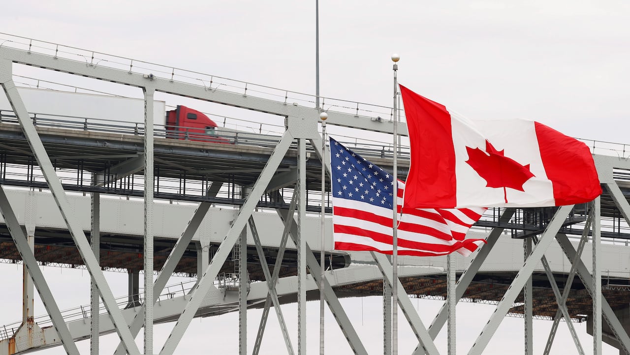 Canadian and American flags in front of a large metal bridge with a semi truck driving