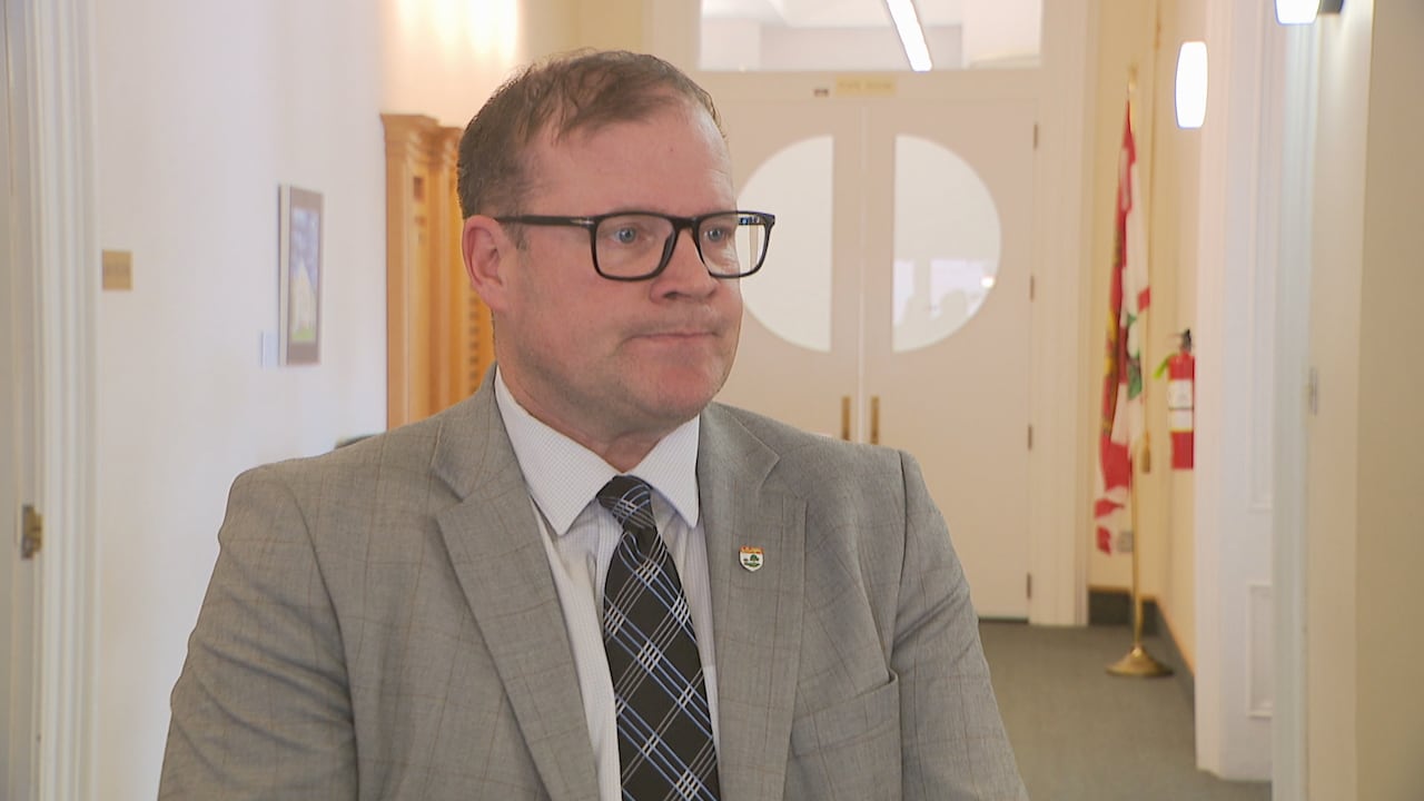 A man in a grey suit and striped tie stands in a white corridor.