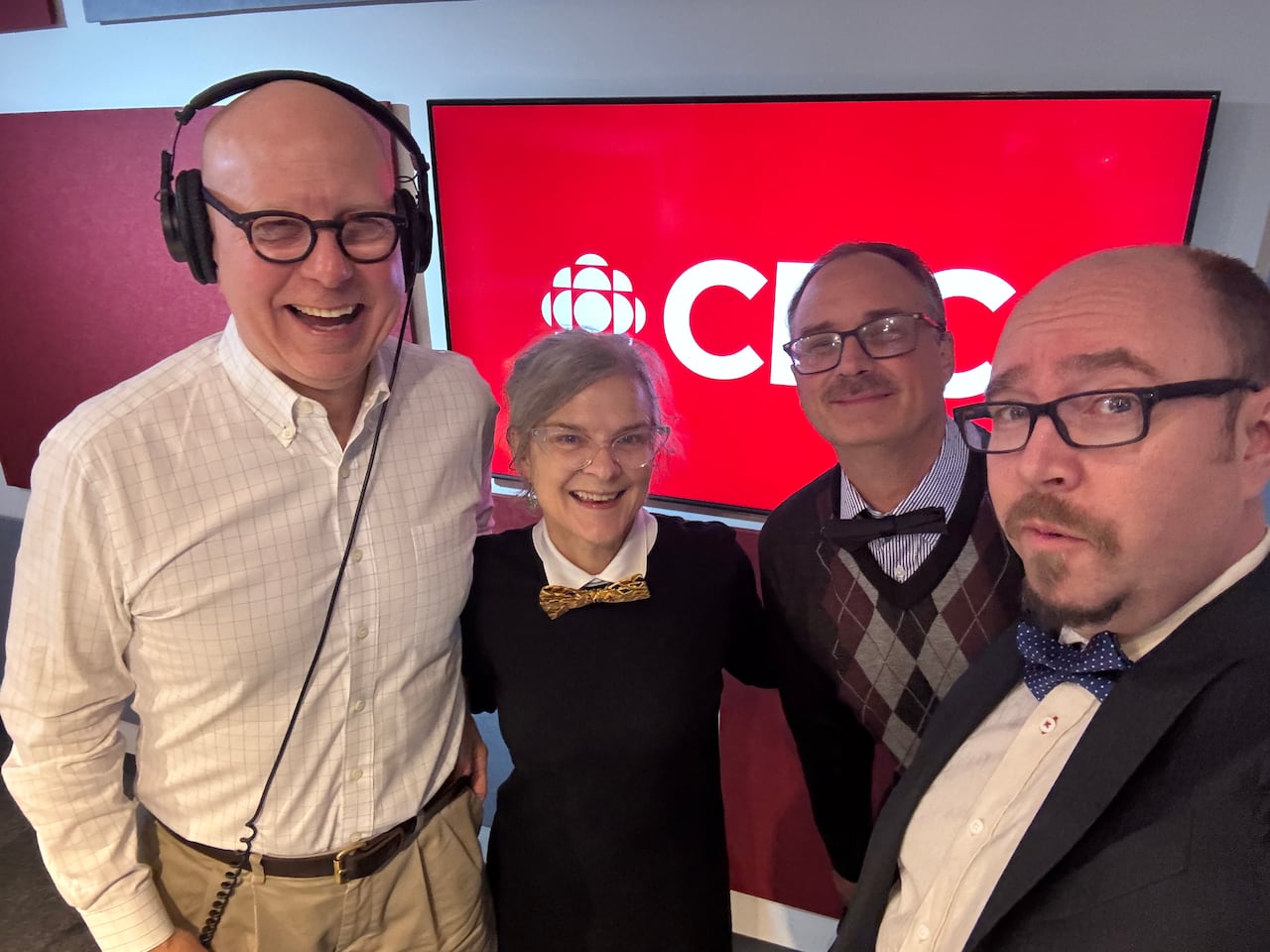 Four people with glasses smile for a photo in front of a TV screen that says CBC on it.