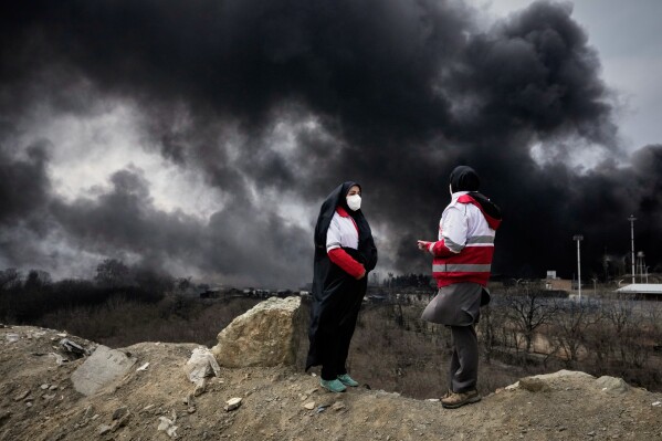 Two women from the Iranian Red Crescent Society stand as a thick plume of smoke from a U.S.-Israeli strike on an oil storage facility late Saturday rises into the sky in Tehran, Iran, Sunday, March 8, 2026. (AP Photo/Vahid Salemi, File)